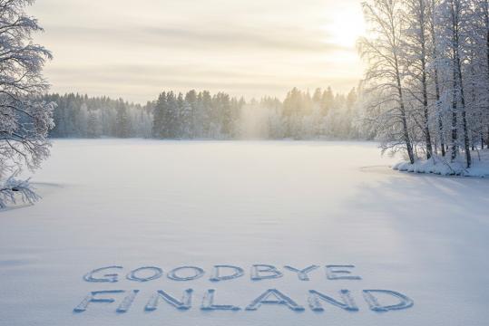 Talvimaisema, jossa on jäätynyt järvi, jota ympäröivät lumen peittämät puut. Aurinko on matalalla taivaalla ja heittää pehmeän kultaisen hehkun sumun läpi. Etualalla tuoreeseen lumeen on huolellisesti painettu teksti ”GOODBYE FINLAND”, joka luo silmiinpistävän kontrastin koskemattomaan valkoiseen pintaan